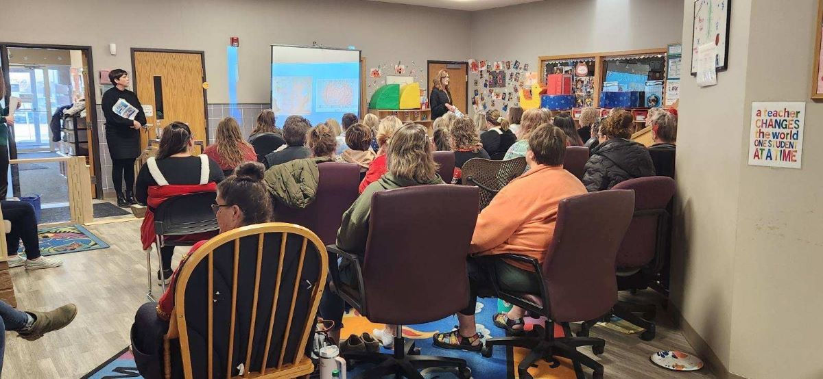 A group of about 20 teachers learning. They are sitting down with their backs to the camera looking at a wall display.