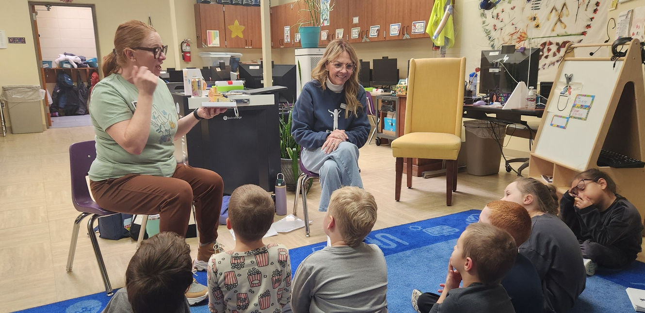 2 SDSD and SD DROP staff teaching sitting on chairs facing a group of elementary age children. Teachers are teaching sign language to then young children