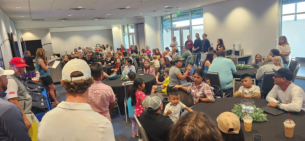Group of elementary school children and adults sitting at large roud tables. The room listeing to a womn at the front of the room reading from a sheet of paper.