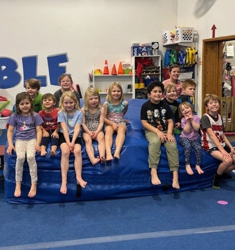 Group of about 10 elementary school children sitting on tumbling matts.