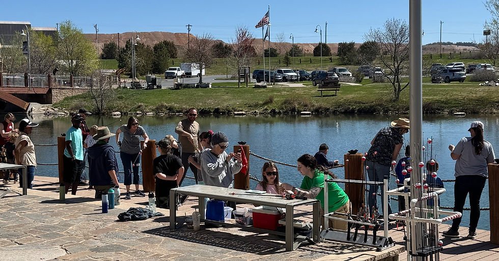 Group of 20 children and adults standing at the side of a lake fishing or putting together fishing rods to go fishing. Spring afternoon with a blue cloud free sky.