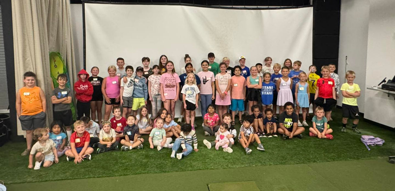 Large group of elementary and middle school age children at an SDSD golf event. The children are sitting and standing in front of white screens on faux green putting grass
